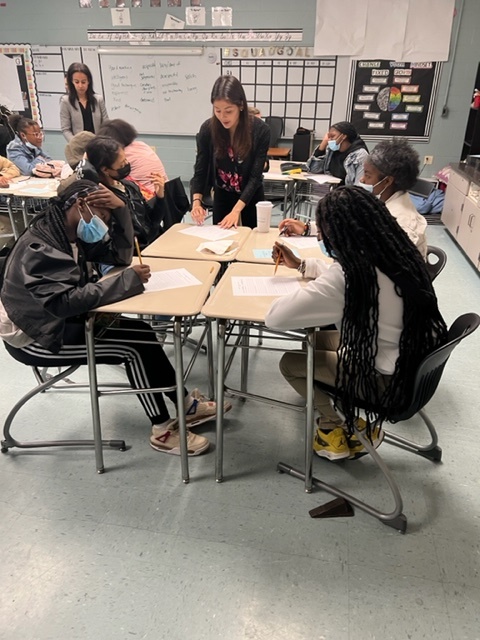 Photo of attorneys from Hunton Andrews Kurth hosting a career fair for Kelly Miller Middle School students. Four students are seated in a classroom wearing masks and looking at sheets of paper while an attorney is slightly leaned over the foursquare of desks and speaking to them